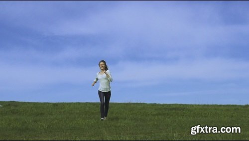 Young happy woman is running and jumping by the camera blue sky and green grass on the background