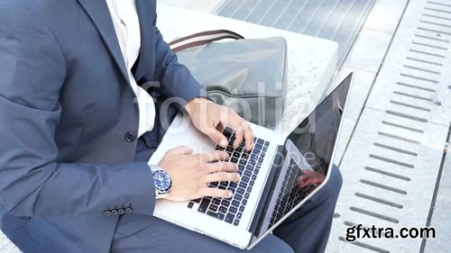 Handsome young man sitting on a stone bench working outdoors in an urban setting typing information onto his laptop computer