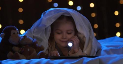 Little Girl Looks at Pictures in a Book, She Holds a Flashlight in Her Hand. Bokeh Background - G98UYQ6