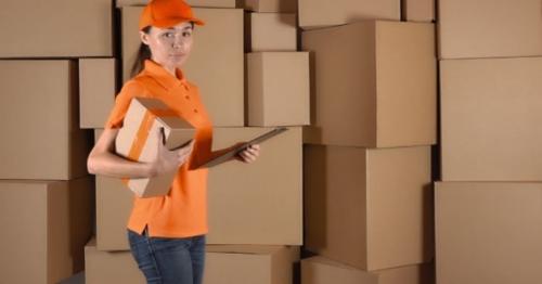 Beautiful Female Courier In Orange Uniform Delivering a Parcel Against Carton Stacks Backround - 4R9H2UP