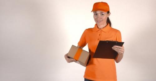 Beautiful Female Courier In Orange Uniform Delivering a Parcel. Light Gray Backround - 65JLVXA