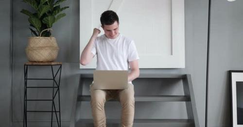 Excited Man Celebrating Win on Laptop while Sitting on stairs - GRE9JVZ