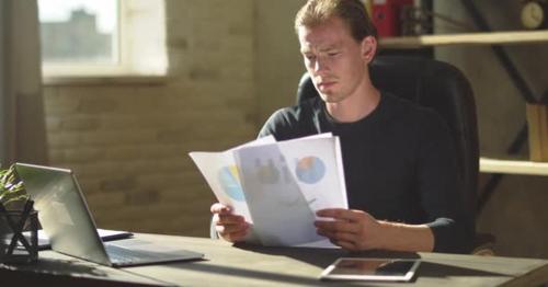 Handsome Concentrated Young Businessman Sitting at the Office Desk and Working with Documents - PXWJRGM