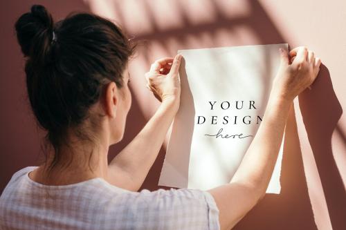 Woman attaching a white poster mockup to a pastel pink wall - 1210126