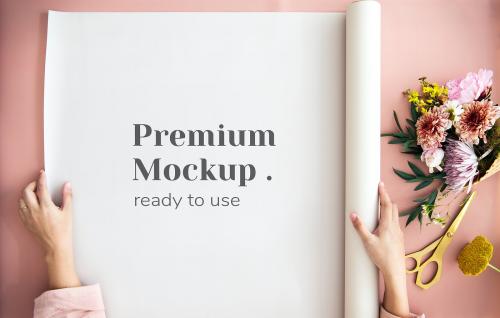 Woman unroll a paper mockup on a pink table by a bouquet of flowers - 1210270