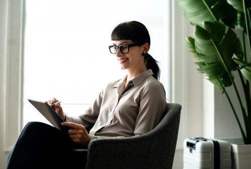 Woman using a stylus with her digital tablet while waiting for her flight in an airport lounge - 1216639