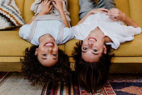 Cheerful women lying upside down on a mustard yellow couch - 1200495