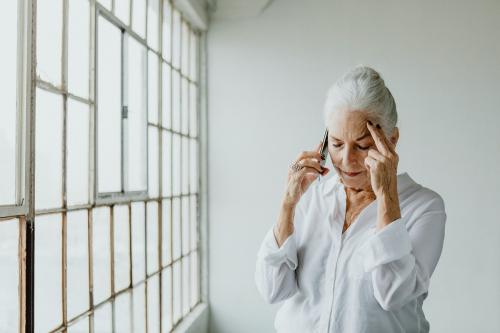 Stressed senior woman talking on a phone by the window in a white room - 1201630